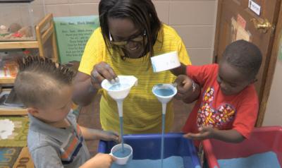 Teacher and children play with funnels at sand table.