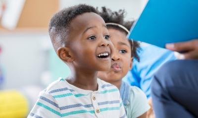 A young boy looking excited and happy as his teacher reads a book.