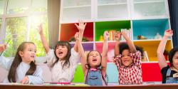 Diverse group of children in a classroom raising their hands in the air!