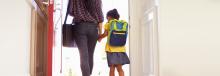 A young girl with a backpack walks through a door with her parent.  Both are seen from behind.