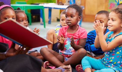 A group of children listens as a teacher reads them a book.